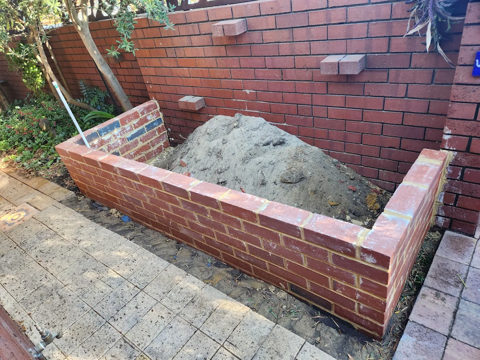 Newly built red brick planter box in a residential backyard, with fresh sand fill inside and existing brick boundary walls behind, showcasing neat brickwork and clean mortar joints.